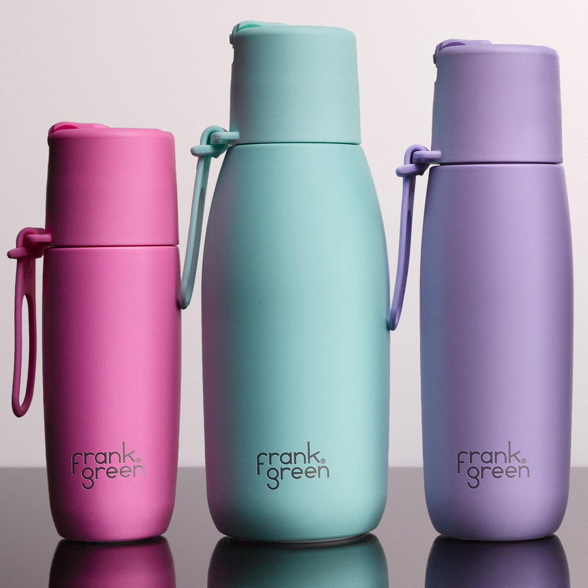 Three Frank Green insulated water bottles in pink, mint green, and lavender purple, arranged side-by-side on a reflective dark surface against a light gray background.