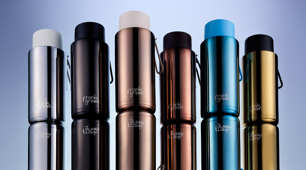 Six metallic travel tumblers with different colors (silver, black, rose gold, bronze, blue, and gold) lined up vertically on a reflective surface against a blue gradient background. The brand name 'frank graham' is printed near the bottom of each tumbler.