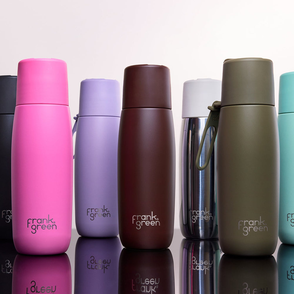A lineup of Frank Green stainless steel water bottles in various colors, including pink, purple, brown, silver, and olive green, arranged on a reflective white surface against a light gray background.