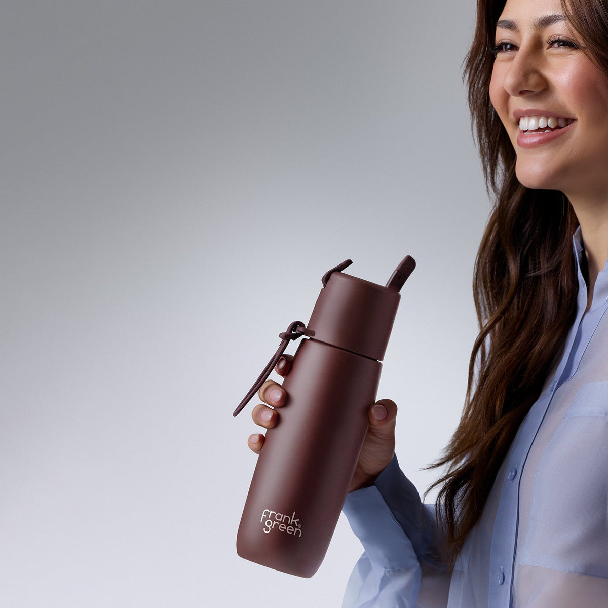 A woman in a light blue shirt holds a matte dark brown insulated water bottle with a straw and a small zipper pull. The bottle has the 'crank & green' logo printed near the bottom.