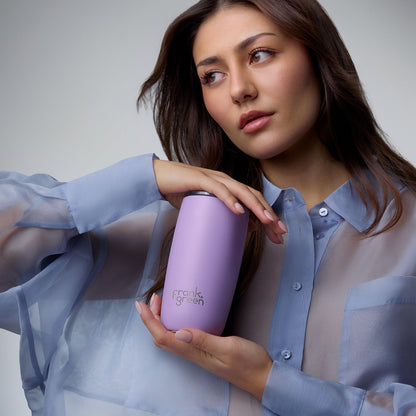 A woman in a sheer light blue button-up shirt holds a lavender purple insulated tumbler with the brand name 'frank's garden' printed on it, against a neutral gray studio background.