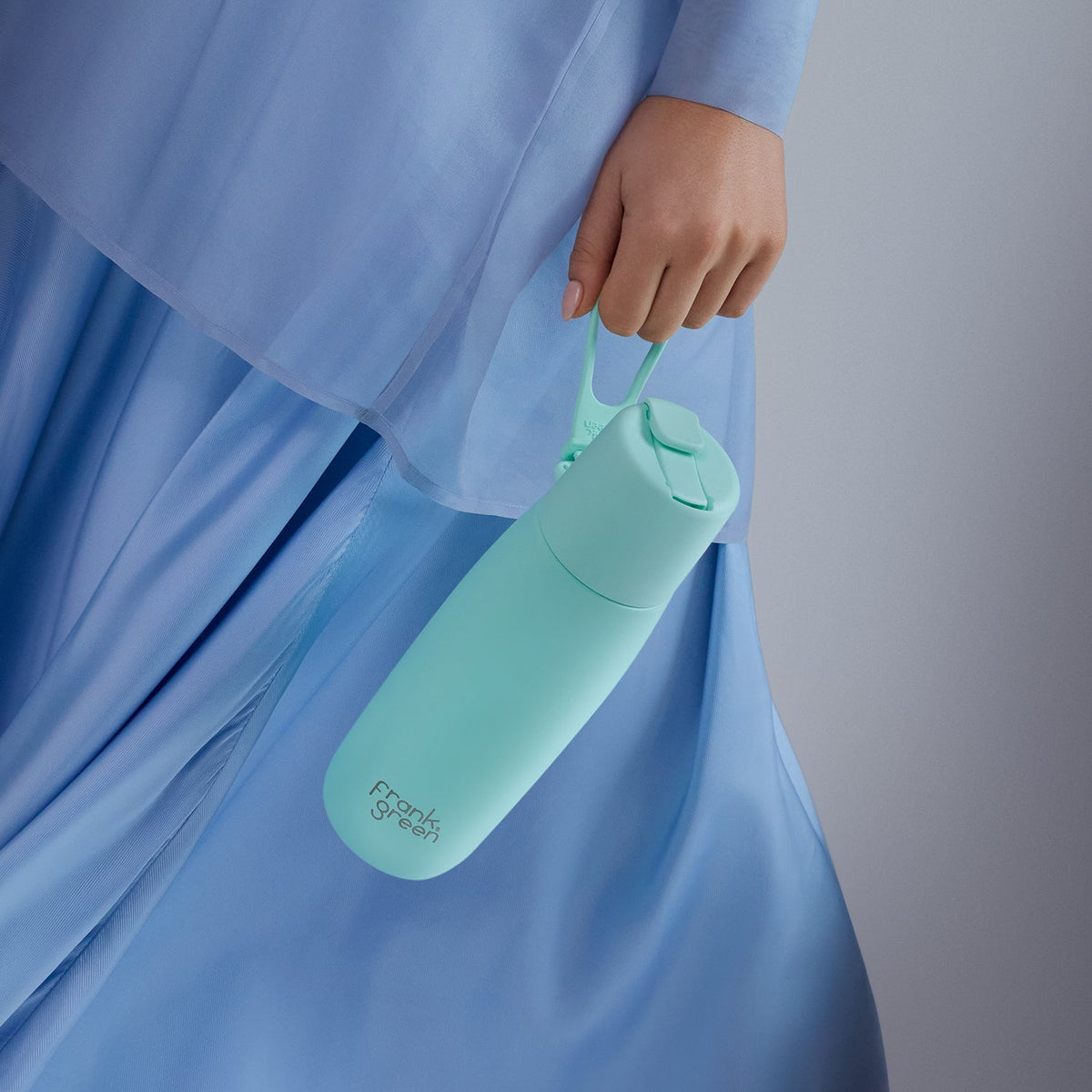 A mint green reusable water bottle with a matching carrying loop, held by a person wearing a light blue dress against a soft gray studio background.