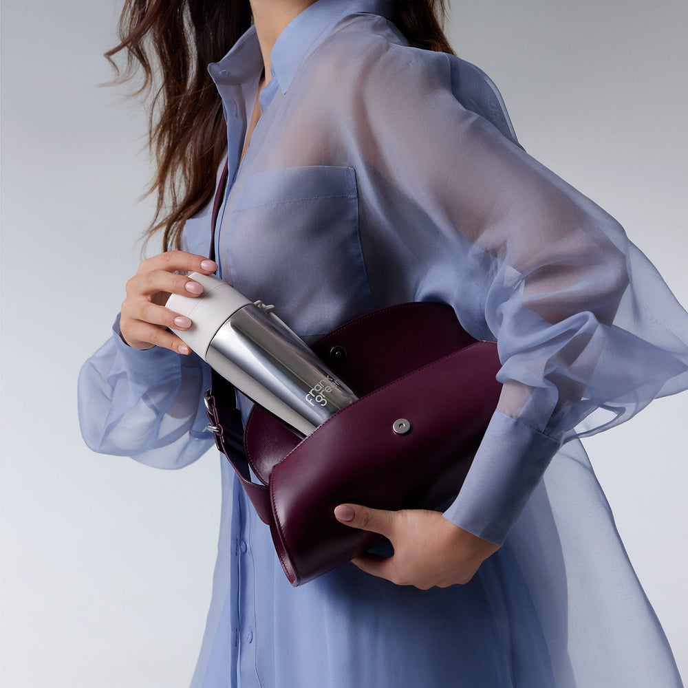 A woman wearing a sheer light blue blouse holds a burgundy leather shoulder bag with a silver metallic bottle inserted into the side pocket, against a clean light gray studio background.