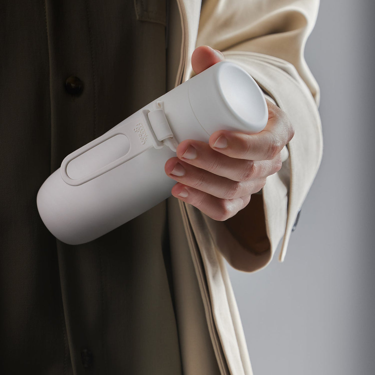 A person in a beige coat holds a white reusable bottle with a flip-top lid. The bottle has a matte finish and a small embossed logo near the cap.