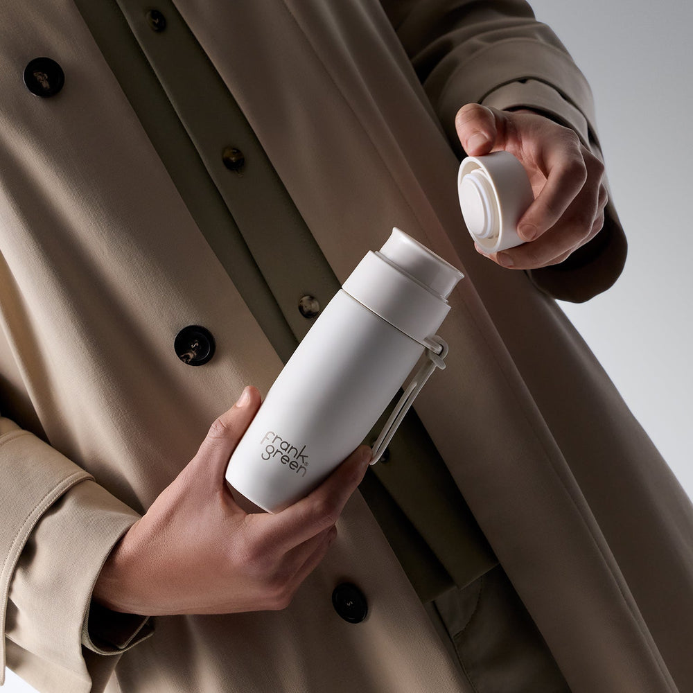 A person in a beige trench coat holds a white vacuum-insulated travel tumbler with a clear lid, showing the brand name 'crankles green' printed on the side. The person is shown from the chest down against a neutral light gray background.