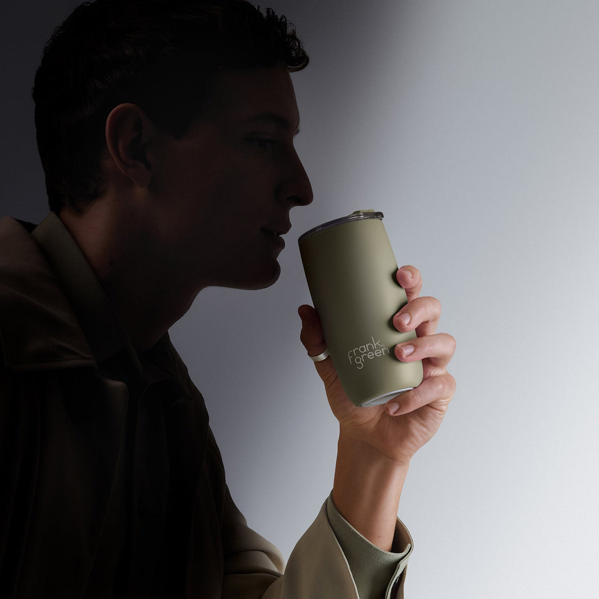A man in profile holds a matte olive green travel tumbler with a silver lid. The tumbler has the text 'crank of west' printed on it.