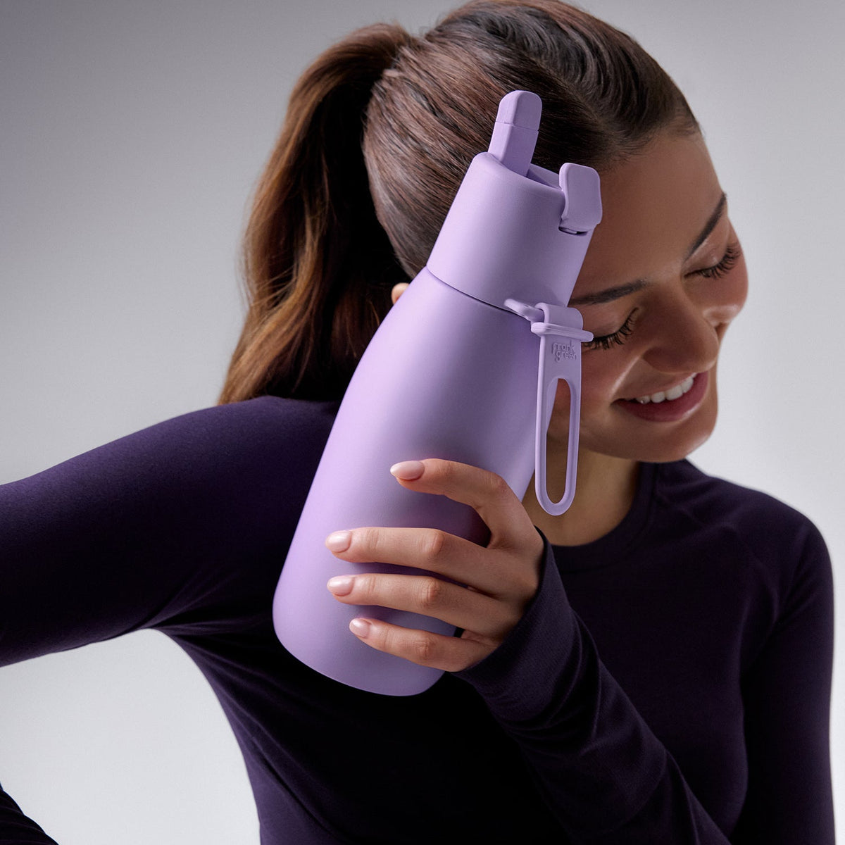 A woman in a dark purple long-sleeve top holds a matte lavender water bottle with a carrying loop and flip spout against a neutral gray studio background.