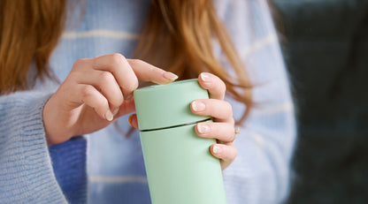 A close-up of a person holding a matte mint green travel tumbler with a matching lid, shown in a lifestyle setting with a cozy background.
